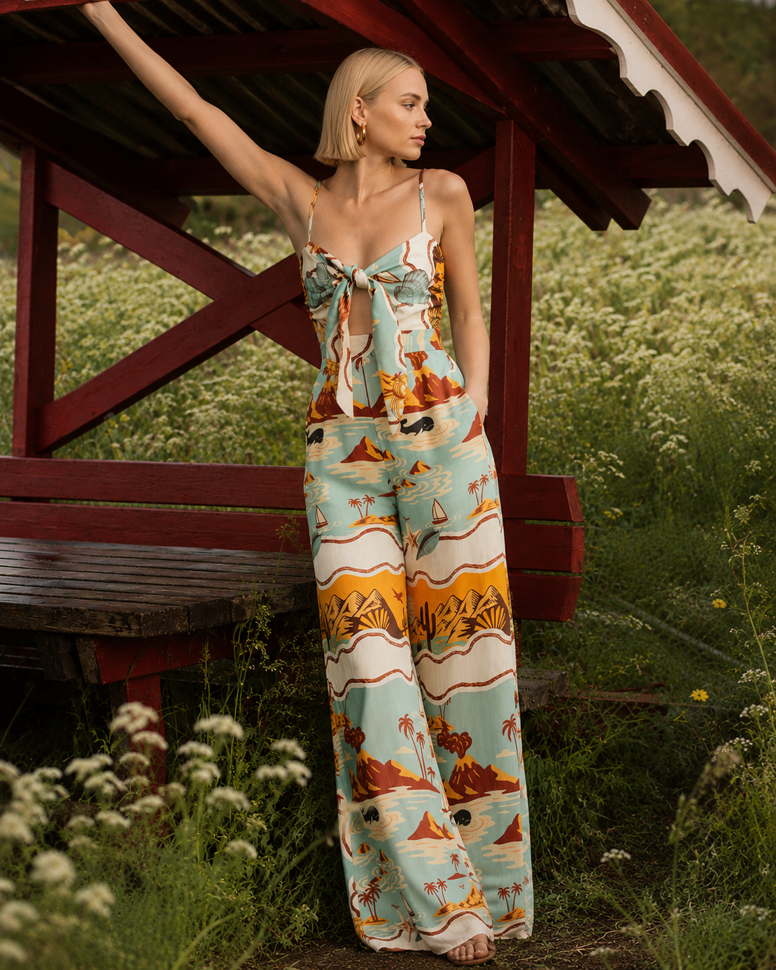 A woman stands barefoot outdoors under a red wooden shelter, wearing a sleeveless wide-leg jumpsuit with a tropical beach print in light blue, orange, beige, and brown. The background features green grass and white wildflowers.